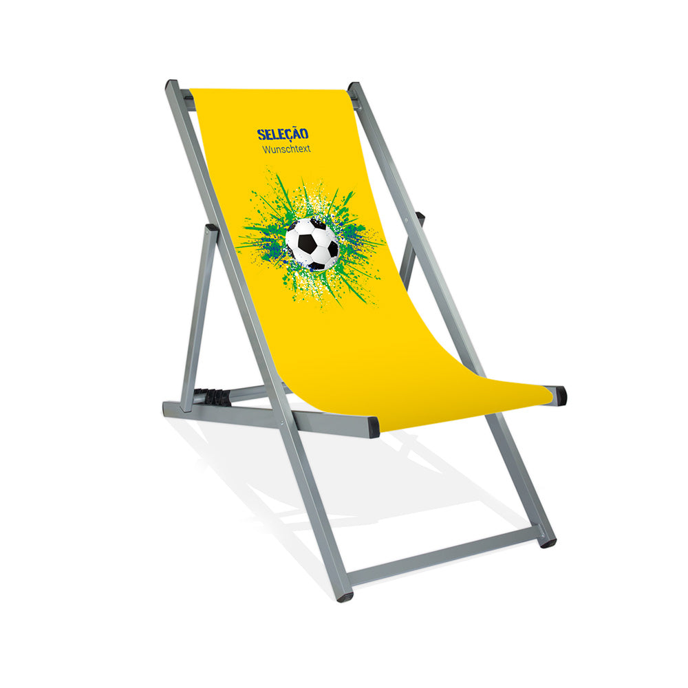 Aluminiumliegestuhl in Silber im Brasilien Design zur WM 2026 mit Fußball auf grün-blau-weißen Farbklecks und personalisierbaren Text oberhalb. Über dem Wunschtext steht in blau "Selecao".
Der Hintergrund ist gelb.