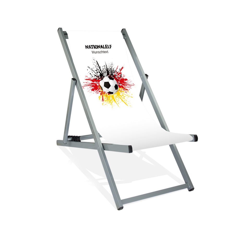 Aluminiumliegestuhl in Silber im Deutschland Design zur WM 2026 mit Fußball auf schwarz-rot-goldenem Farbklecks und personalisierbaren Text oberhalb. Über dem Wunschtext steht "Nationalelf"