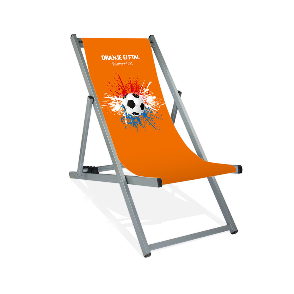Aluminiumliegestuhl in Silber im Niederlande Design zur WM 2026 mit Fußball auf rot-weiß-blauen Farbklecks und personalisierbaren Text oberhalb. Über dem Wunschtext steht "Oranje Elftal". Der Hintergrund ist orange.