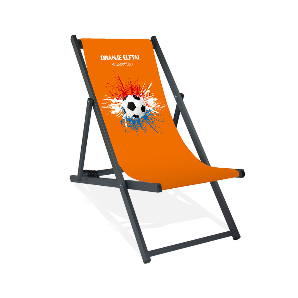 Aluminiumliegestuhl in Schwarz im Niederlande Design zur WM 2026 mit Fußball auf rot-weiß-blauen Farbklecks und personalisierbaren Text oberhalb. Über dem Wunschtext steht "Oranje Elftal". Der Hintergrund ist orange.