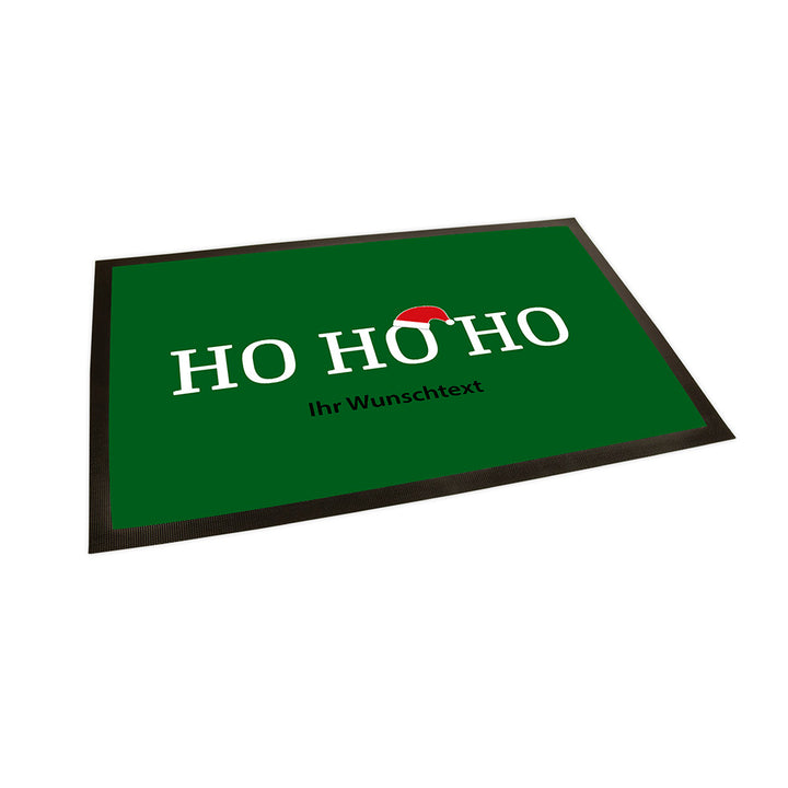 Der Hintergrund der Fußmatte ist in einem Tannengrün gehalten. In der Mitte steht groß und in Weiß geschrieben "Ho Ho Ho". Über dem zweiten "O" ist eine Weihnachtsmannmütze angebracht.
Unter dem Text steht klein und in dunkler Farbe geschrieben "Ihr Wunschtext".