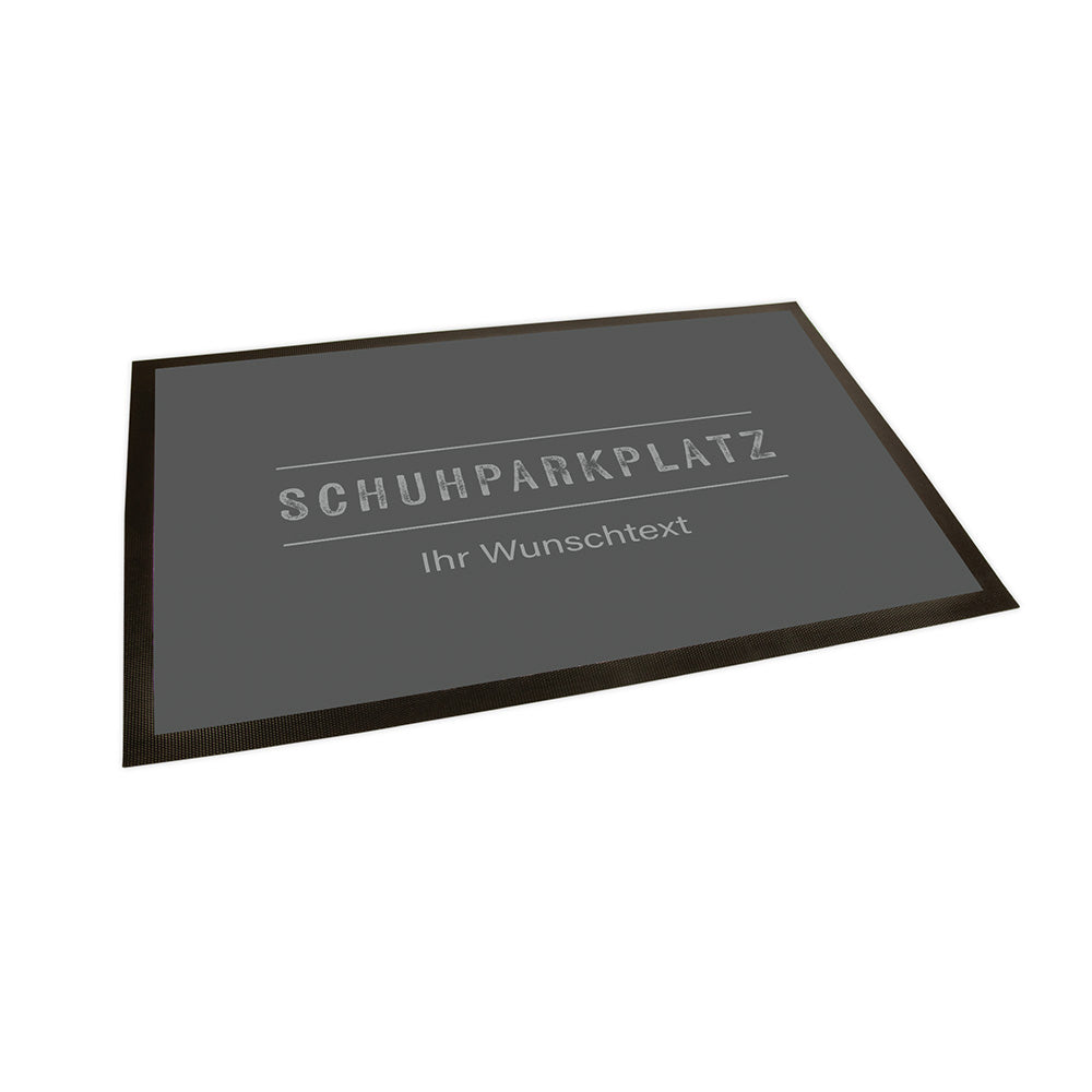 Der Hintergrund der Fußmatte ist in einem dunklen Grau gehalten. In einem hellen Grau steht mittig "Schuhparkplatz" welcher von zwei horizontalen Linien oben und unten eingerahmt wird. Unter der unteren Linie steht als Text in einer anderen Schriftart "Ihr Wunschtext".