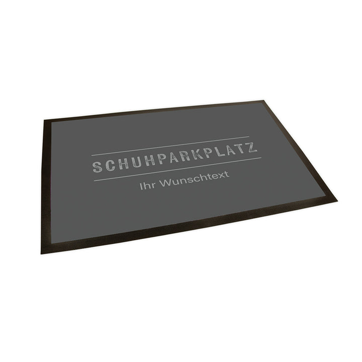 Der Hintergrund der Fußmatte ist in einem dunklen Grau gehalten. In einem hellen Grau steht mittig "Schuhparkplatz" welcher von zwei horizontalen Linien oben und unten eingerahmt wird. Unter der unteren Linie steht als Text in einer anderen Schriftart "Ihr Wunschtext".