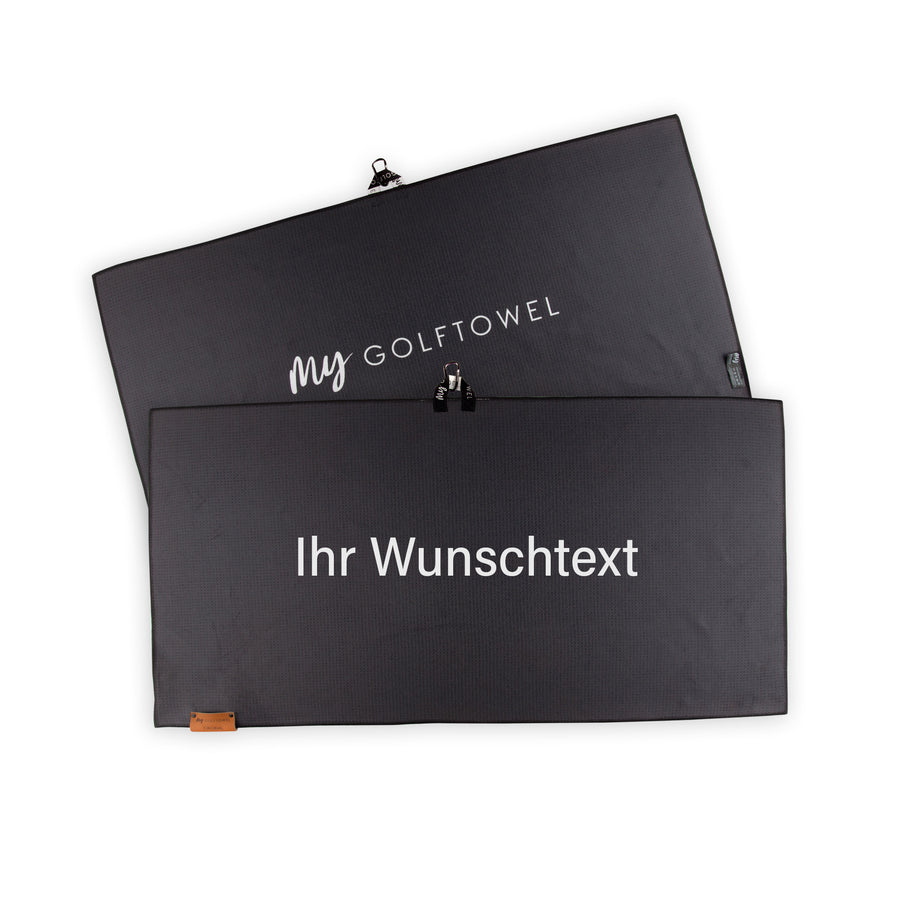 Golftowel Large in Schwarz. Auf dem Golftowel steht Mittig in einer weißen, serifenlosen Schrift geschrieben "Ihr Wunschtext". Die Rückseite des Golftowels ist im gleichen Schwarz, wie die Vorderseite dargestellt. Hier steht mittig der Schriftzug "MyGolftowel". Im unteren Bereich befindet sich ein braunes Lederpatch und oben ist eine Schlaufe und Karabiner angebracht.