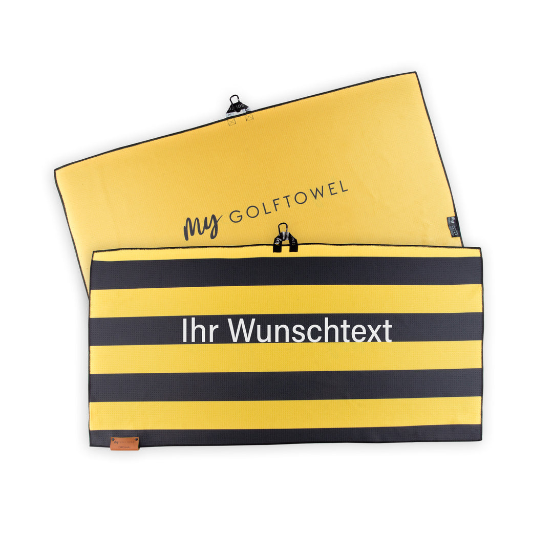 Golftowel Large mit einem horizontal gestreiften Muster Gelb-Schwarz. Auf dem Golftowel steht Mittig in einer weißen, serifenlosen Schrift geschrieben "Ihr Wunschtext". Die Rückseite des Golftowels ist in Gelb, dargestellt. Hier steht mittig der schwarze Schriftzug "MyGolftowel". Im unteren Bereich befindet sich ein braunes Lederpatch und oben ist eine Schlaufe und Karabiner angebracht.